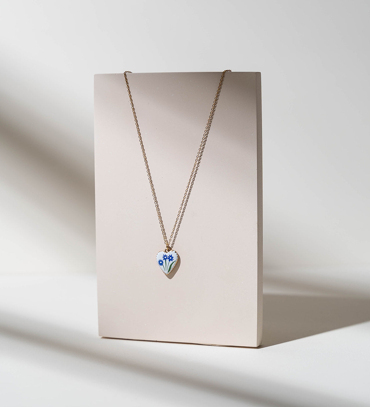 A delicate gold chain necklace with a small heart-shaped pendant featuring blue and white floral details, displayed on a minimalist beige card against a warm, sunlit neutral background.