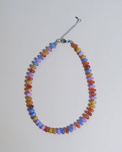 A beaded necklace made of small, faceted beads in multiple colors including orange, yellow, purple, blue, and red, arranged in an oval shape with a silver-tone chain extender and clasp on a light gray background.