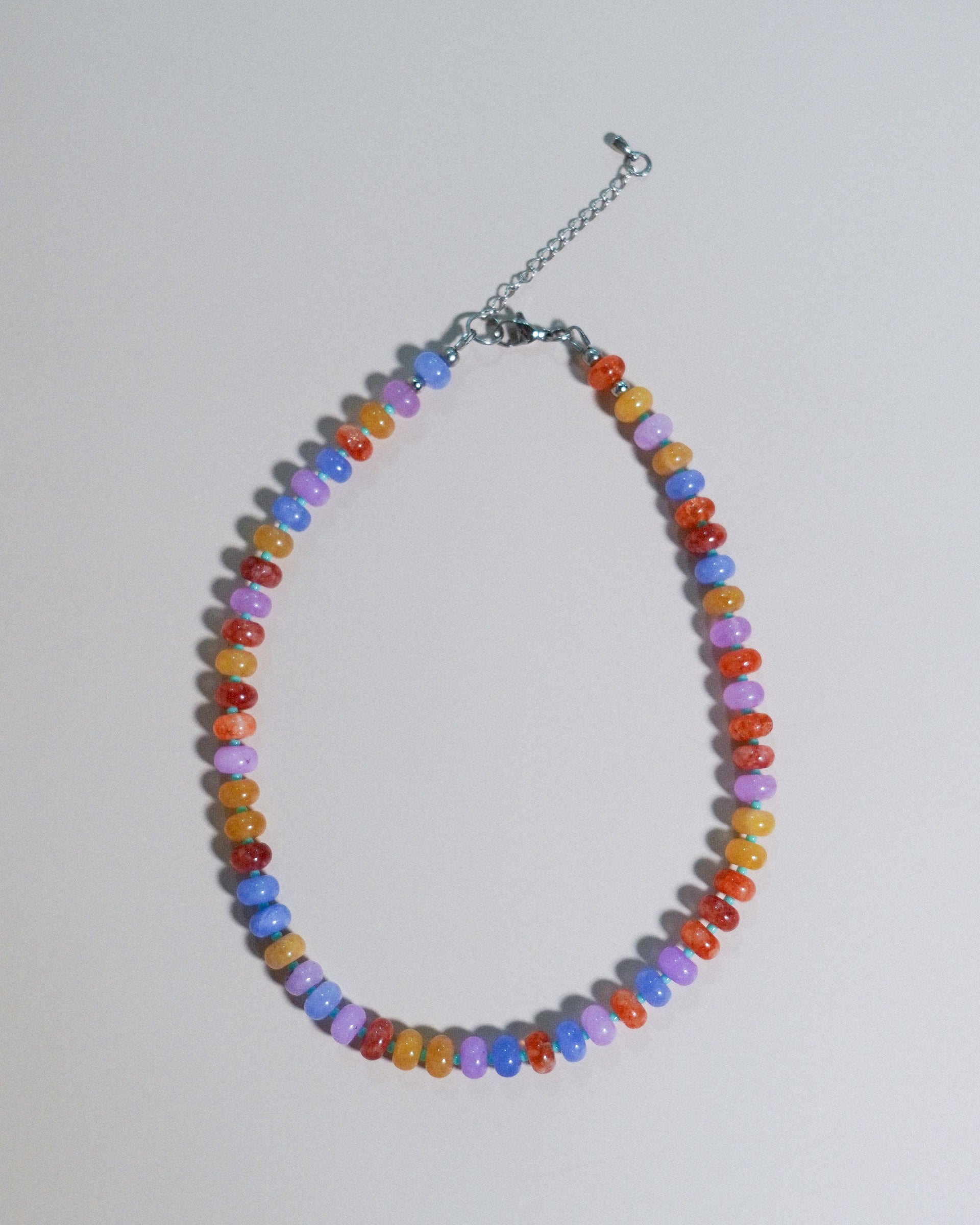 A beaded necklace made of small, faceted beads in multiple colors including orange, yellow, purple, blue, and red, arranged in an oval shape with a silver-tone chain extender and clasp on a light gray background.