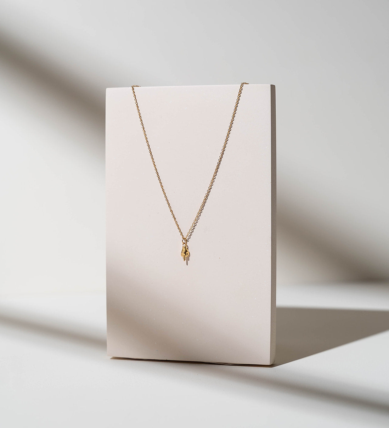 A delicate gold chain necklace with a small textured gold pendant, displayed on a minimalist cream-colored rectangular stand against a warm, sunlit off-white background.
