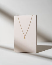 A delicate gold chain necklace with a small textured gold pendant, displayed on a minimalist cream-colored rectangular stand against a warm, sunlit off-white background.