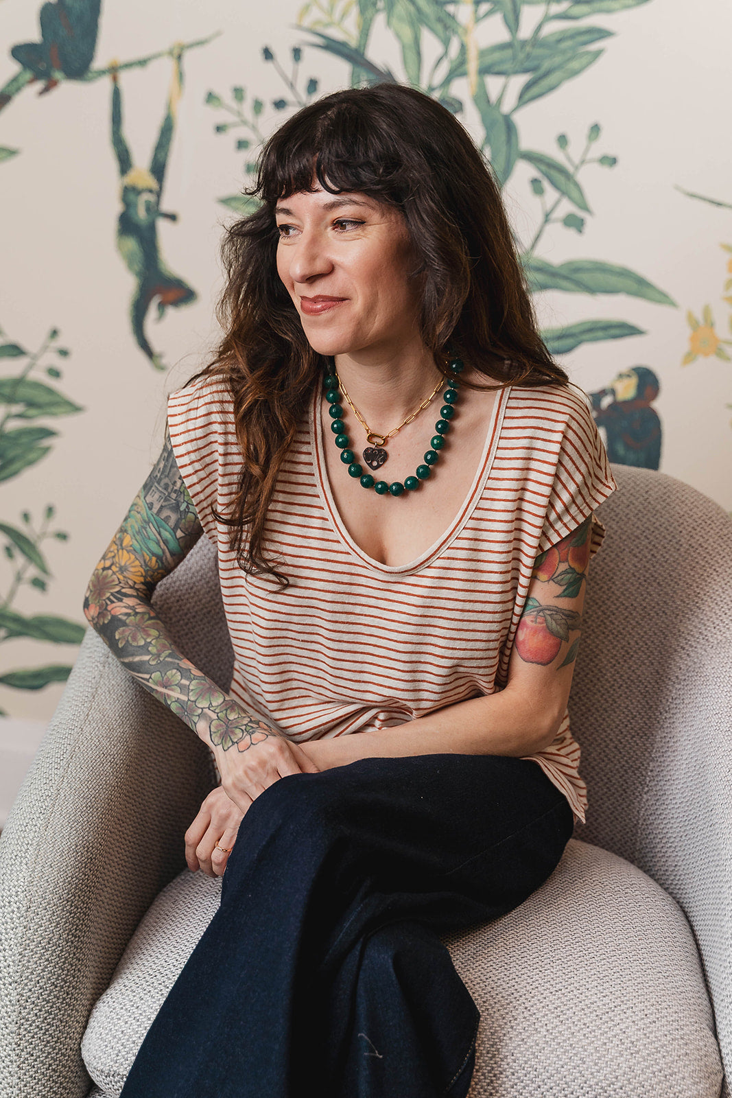Woman with tattoos sitting on a chair in front of a decorative wall with plant patterns.