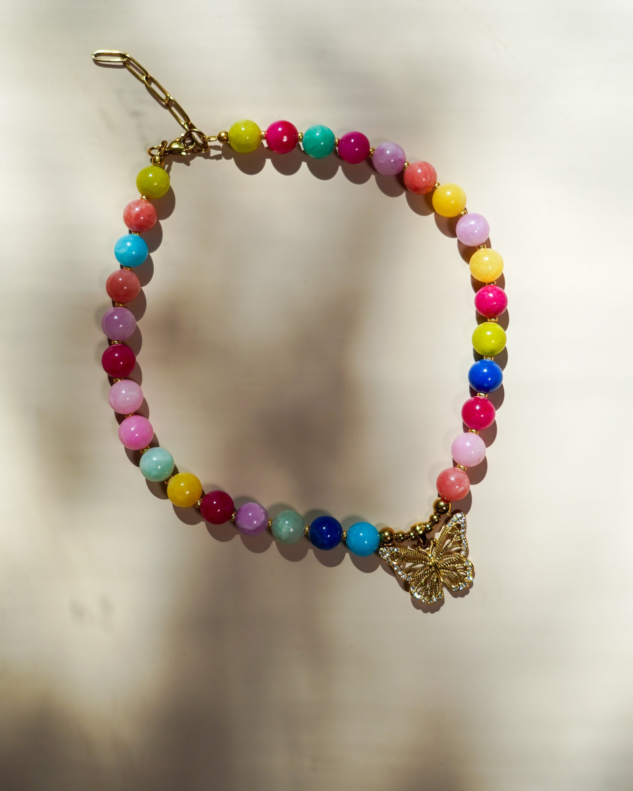A beaded necklace with multicolored round beads and a gold butterfly charm, displayed on a light beige background with soft shadows.