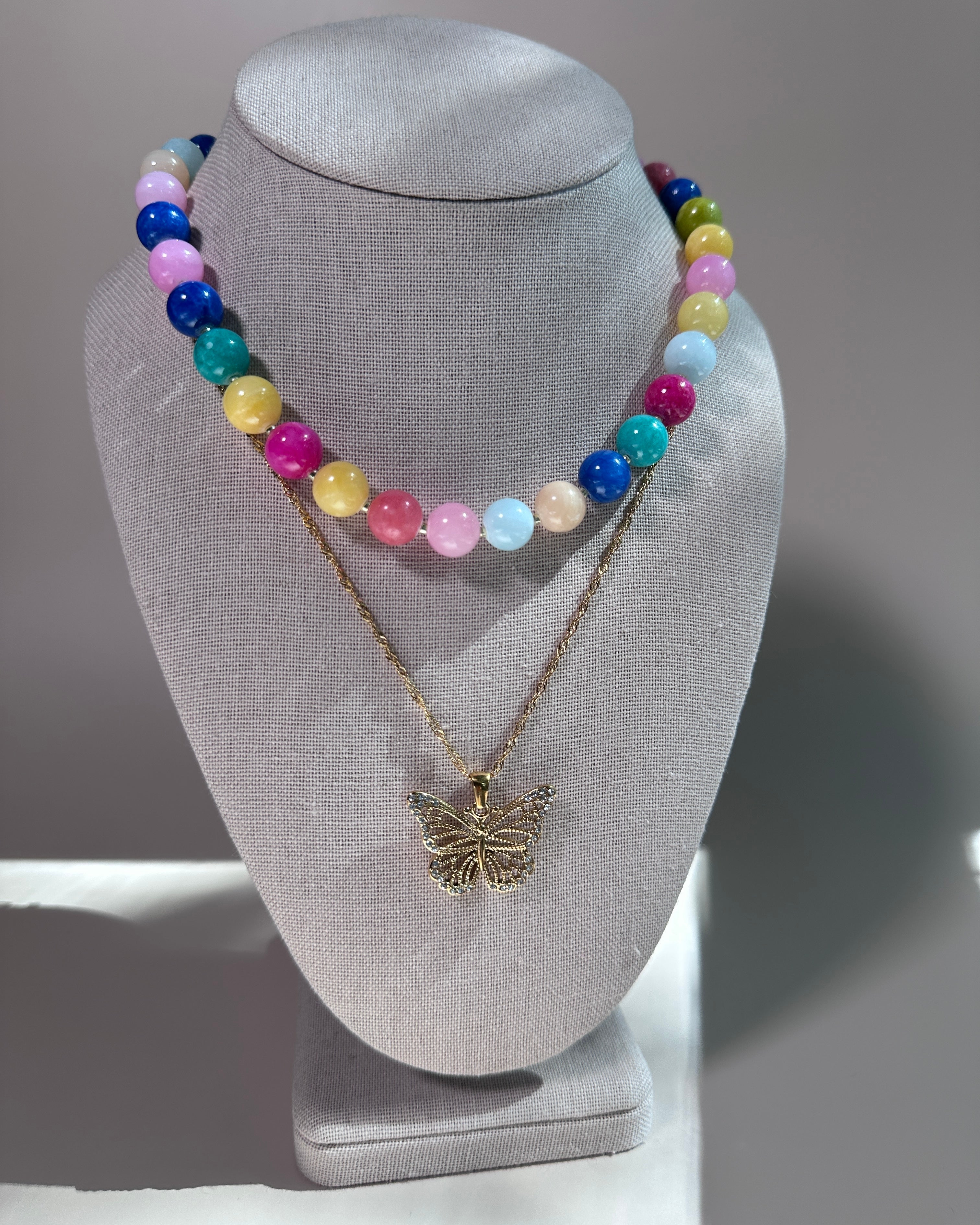 A two-layer necklace displayed on a gray fabric jewelry bust. The top layer features a strand of colorful round beads in pink, blue, yellow, green, and white.