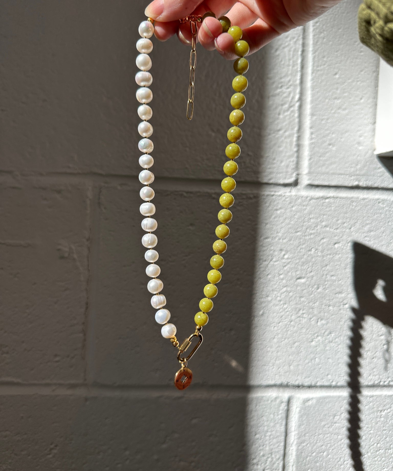 Necklace with green and white beads held by a hand against a tiled wall.