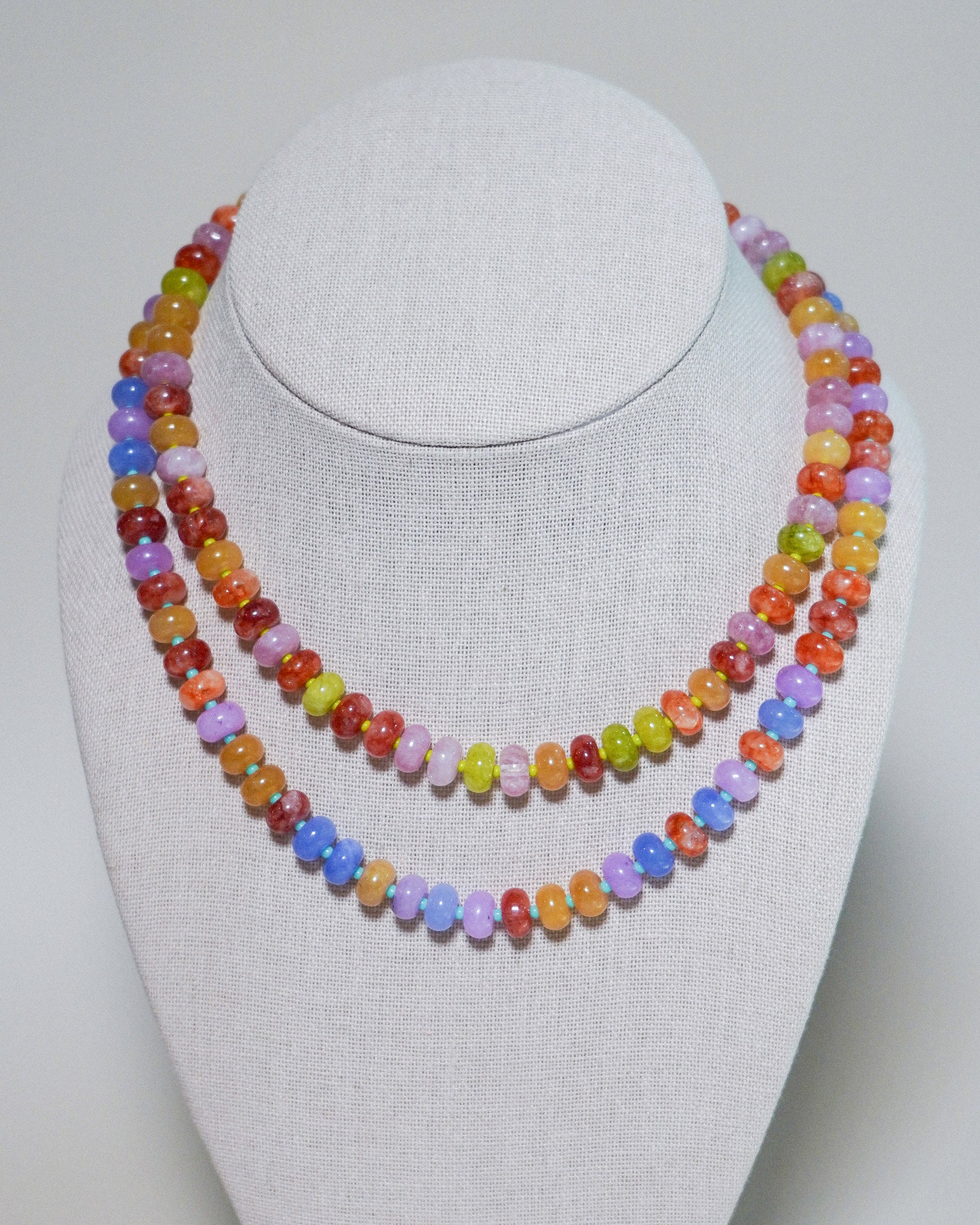 A double-strand beaded necklace displayed on a white fabric-covered jewelry bust. The necklace features small, polished, oval-shaped beads in a variety of bright colors including orange, yellow, green, purple, pink, and blue.
