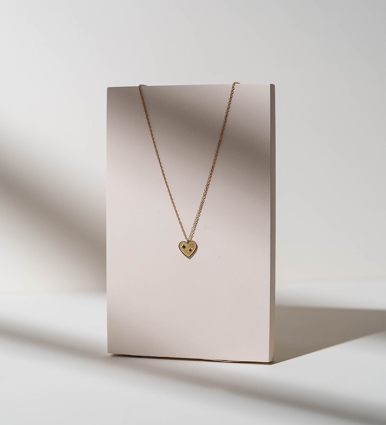 A delicate silver chain necklace with a small teardrop-shaped pendant, displayed on a minimalist beige rectangular stand against a light gray background with soft shadows.