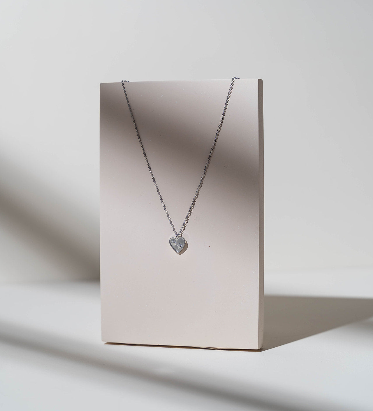 A silver heart pendant necklace displayed on a vertical beige card against a white background with soft shadows.