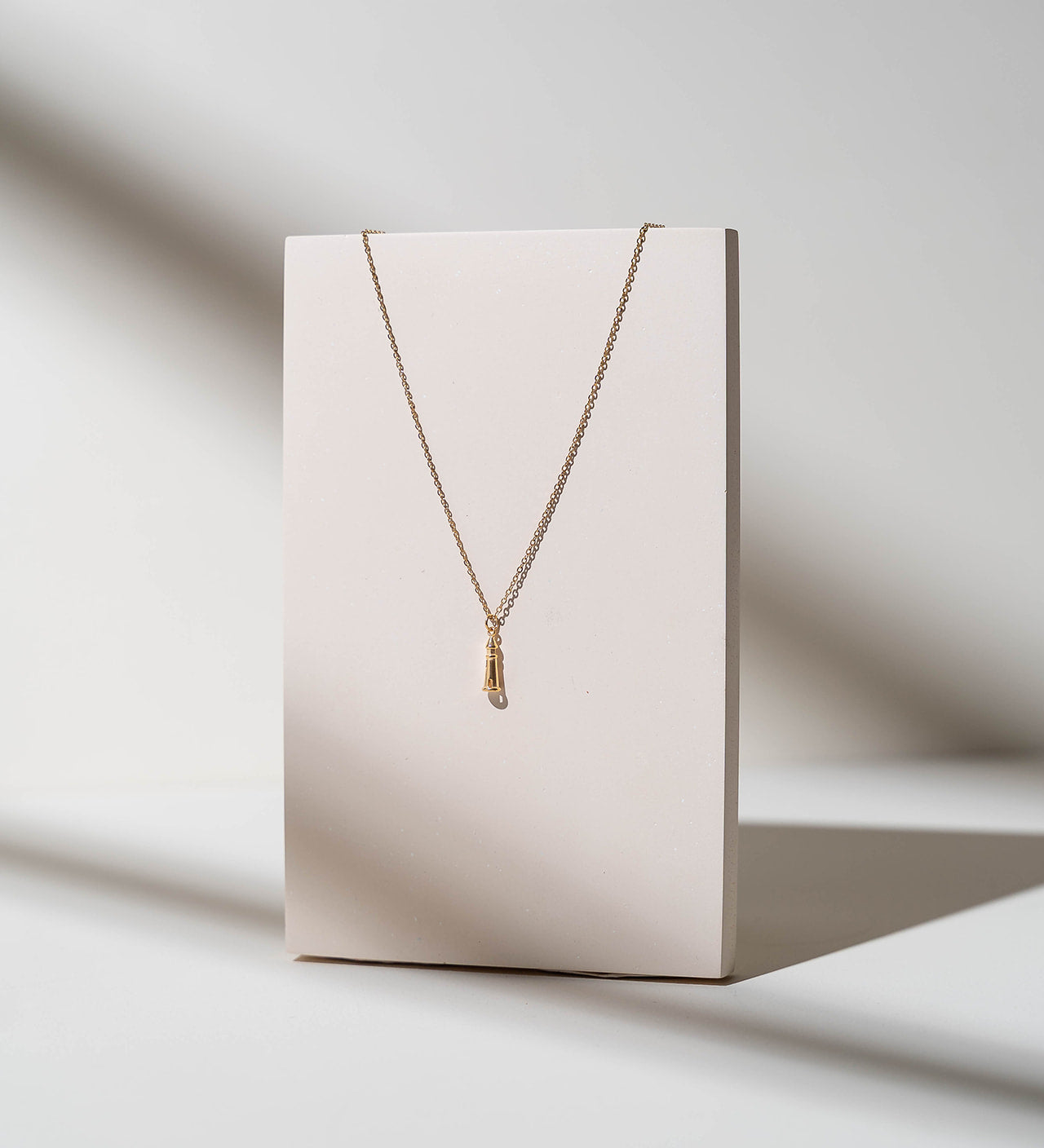 A delicate gold chain necklace with a small textured gold pendant, displayed on a minimalist cream-colored rectangular stand against a warm, sunlit off-white background.