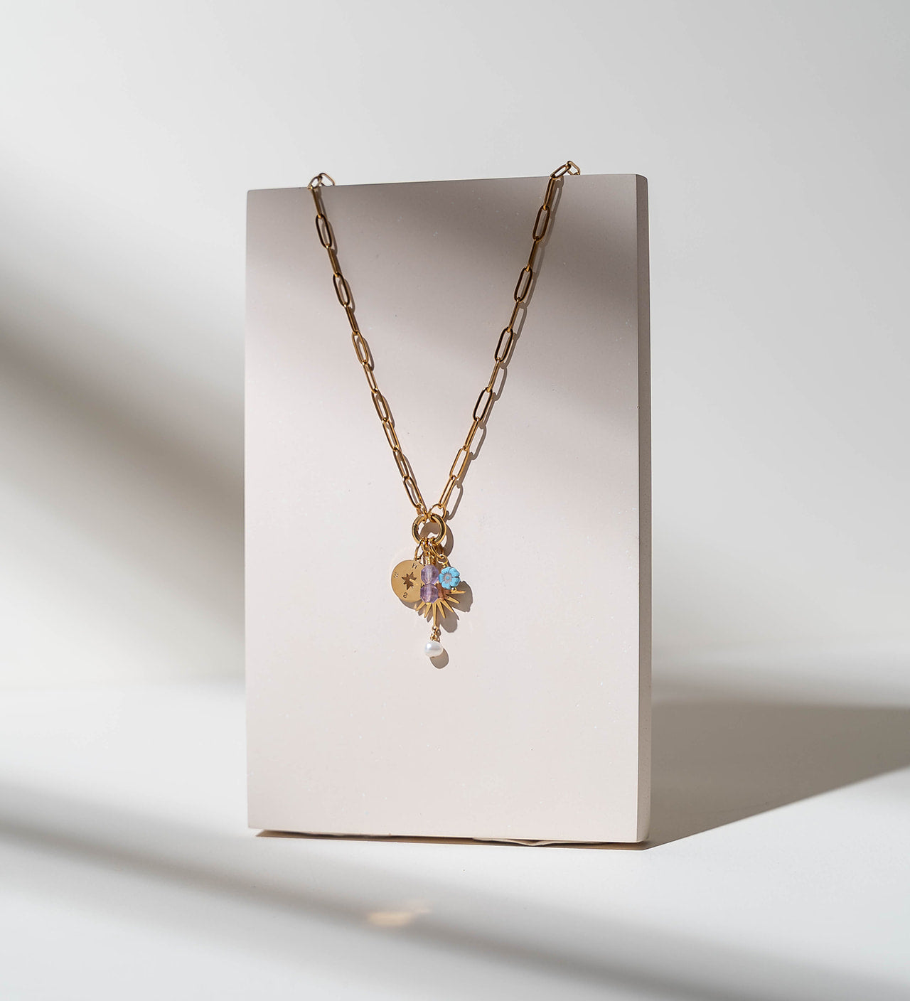 A gold chain necklace with a heart-shaped charm and a small iridescent flower pendant, displayed on a minimalist beige rectangular stand against a white background with soft shadows.