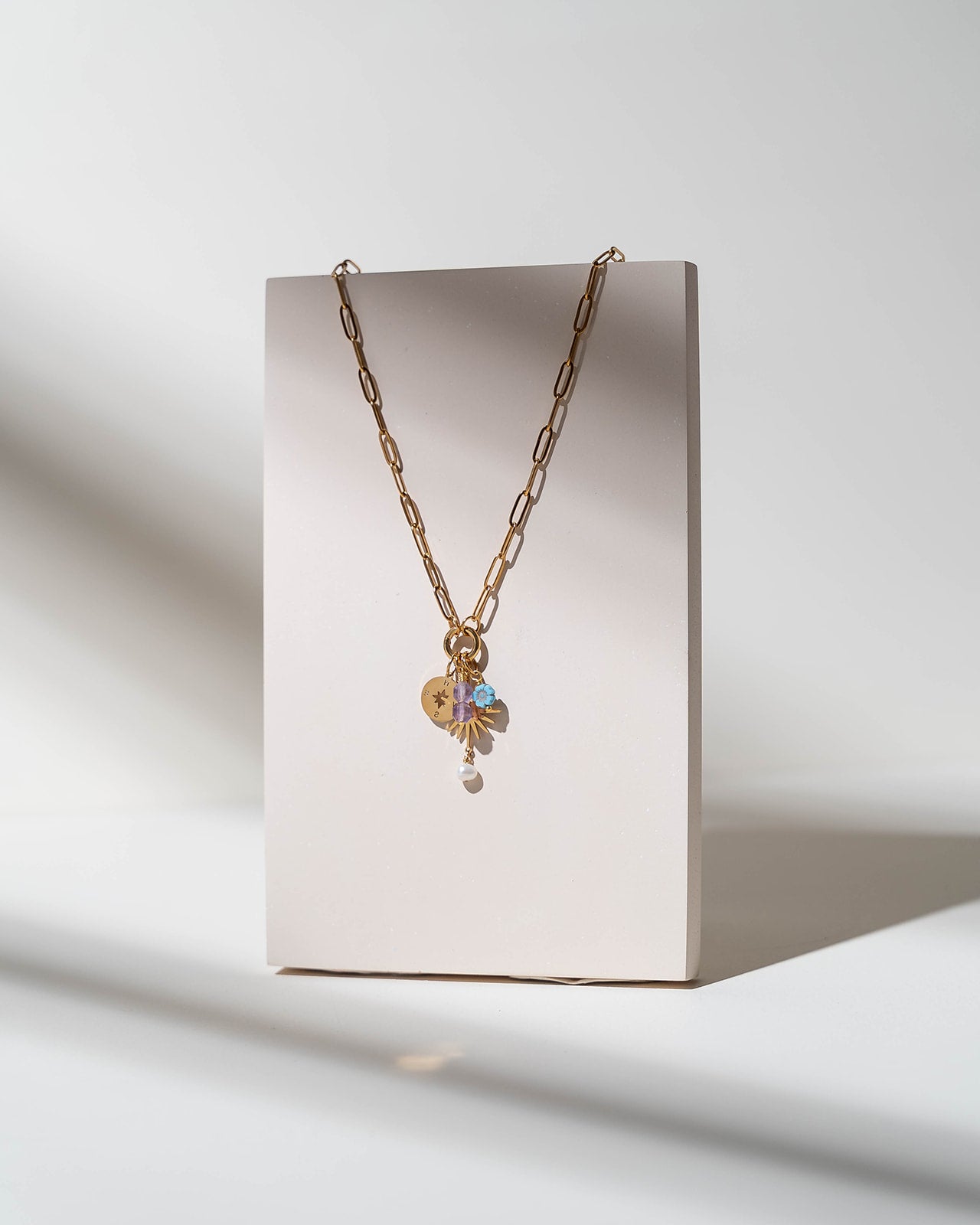 A gold chain necklace with a heart-shaped charm and a small iridescent flower pendant, displayed on a minimalist beige rectangular stand against a white background with soft shadows.