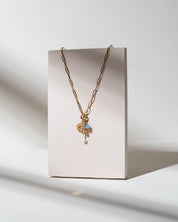 A gold chain necklace with a heart-shaped charm and a small iridescent flower pendant, displayed on a minimalist beige rectangular stand against a white background with soft shadows.
