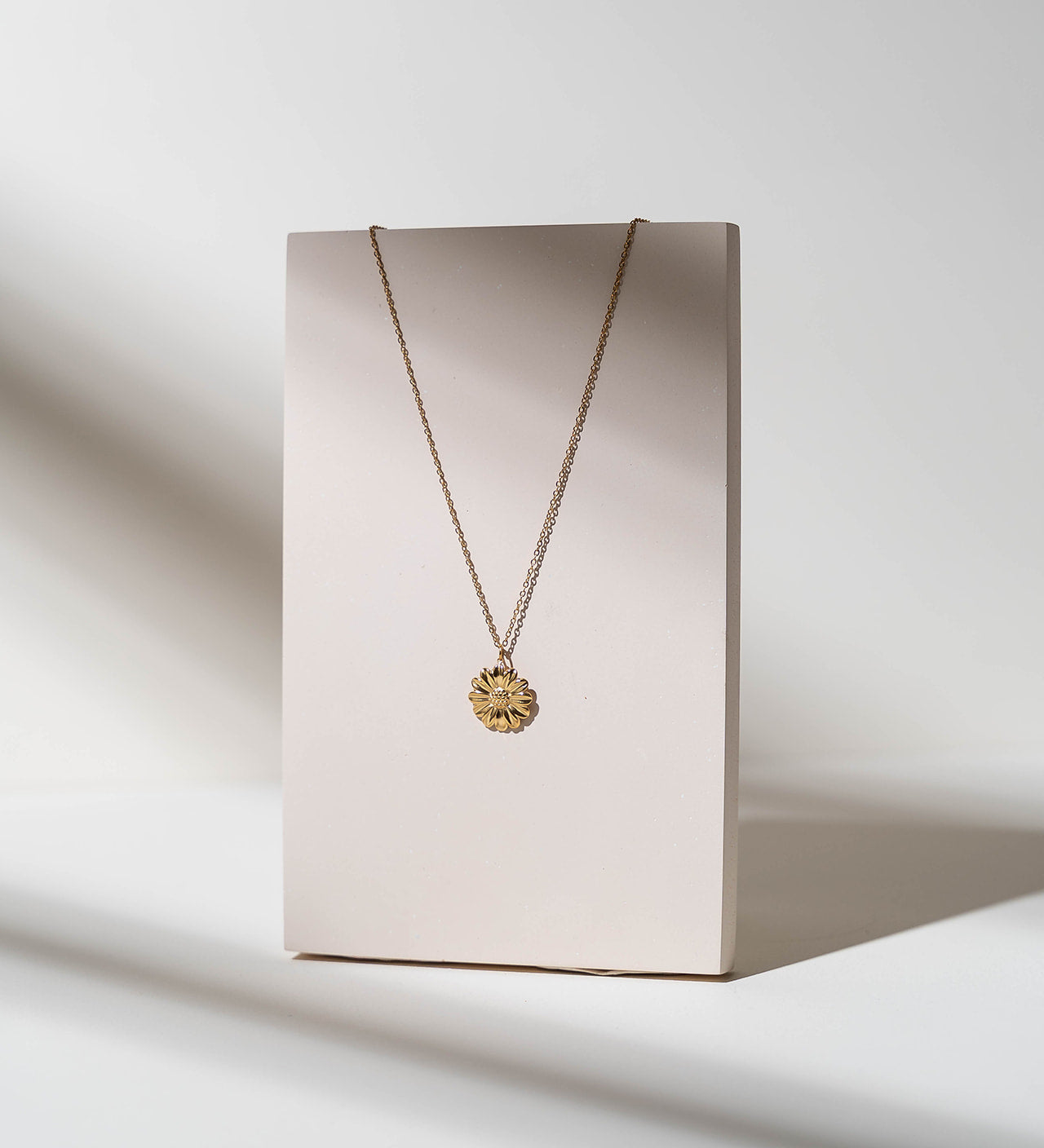 a gold sunflower pendant charm on a chain hanging off a beige pedestal 