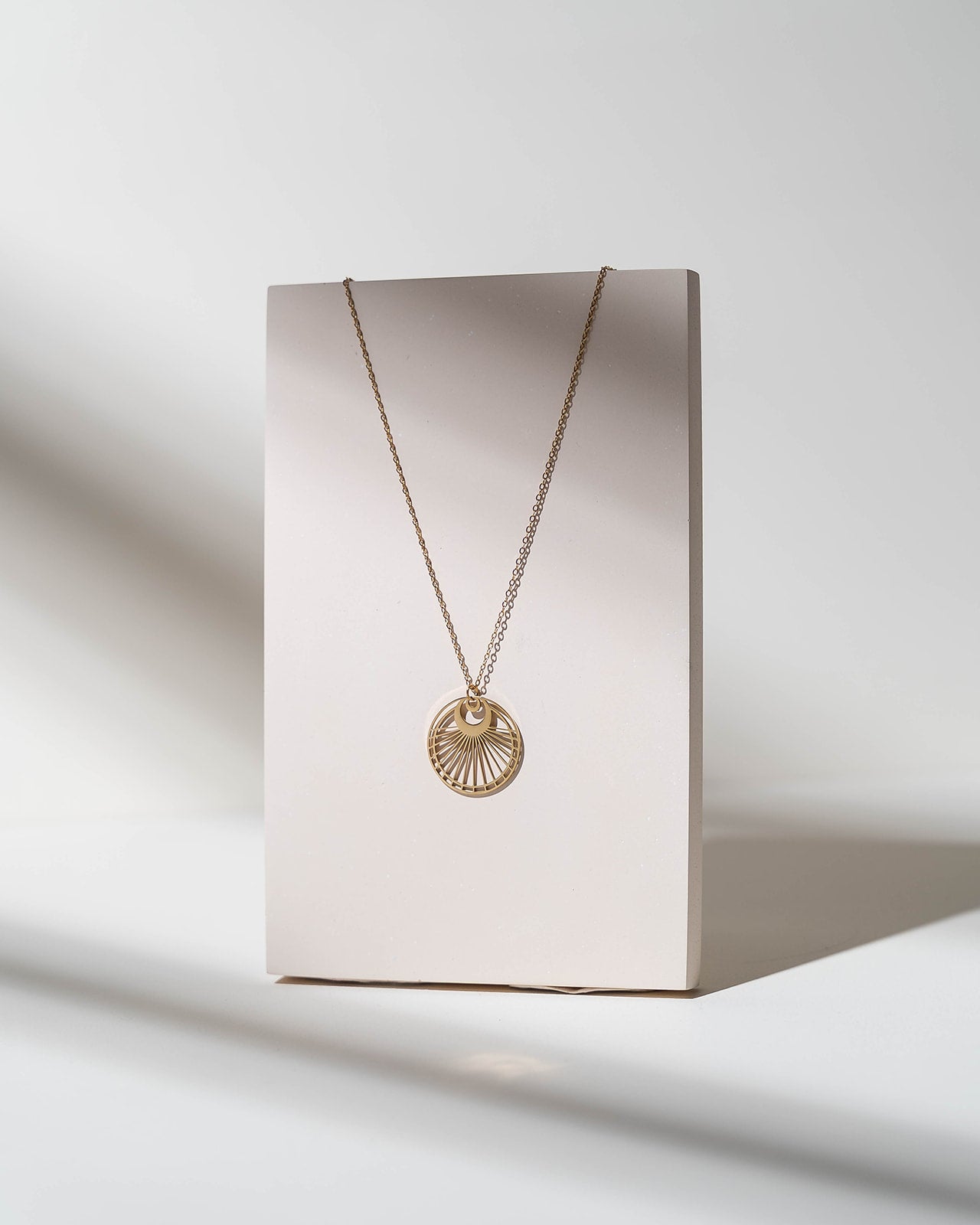 A delicate gold chain necklace with a sunburst pendant displayed on a minimalist beige card, set against a clean white background with soft shadows.