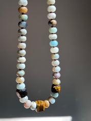 A beaded necklace made of polished gemstone beads in various colors including light blue, white, brown, black, and pink, hanging against a dark gray background.