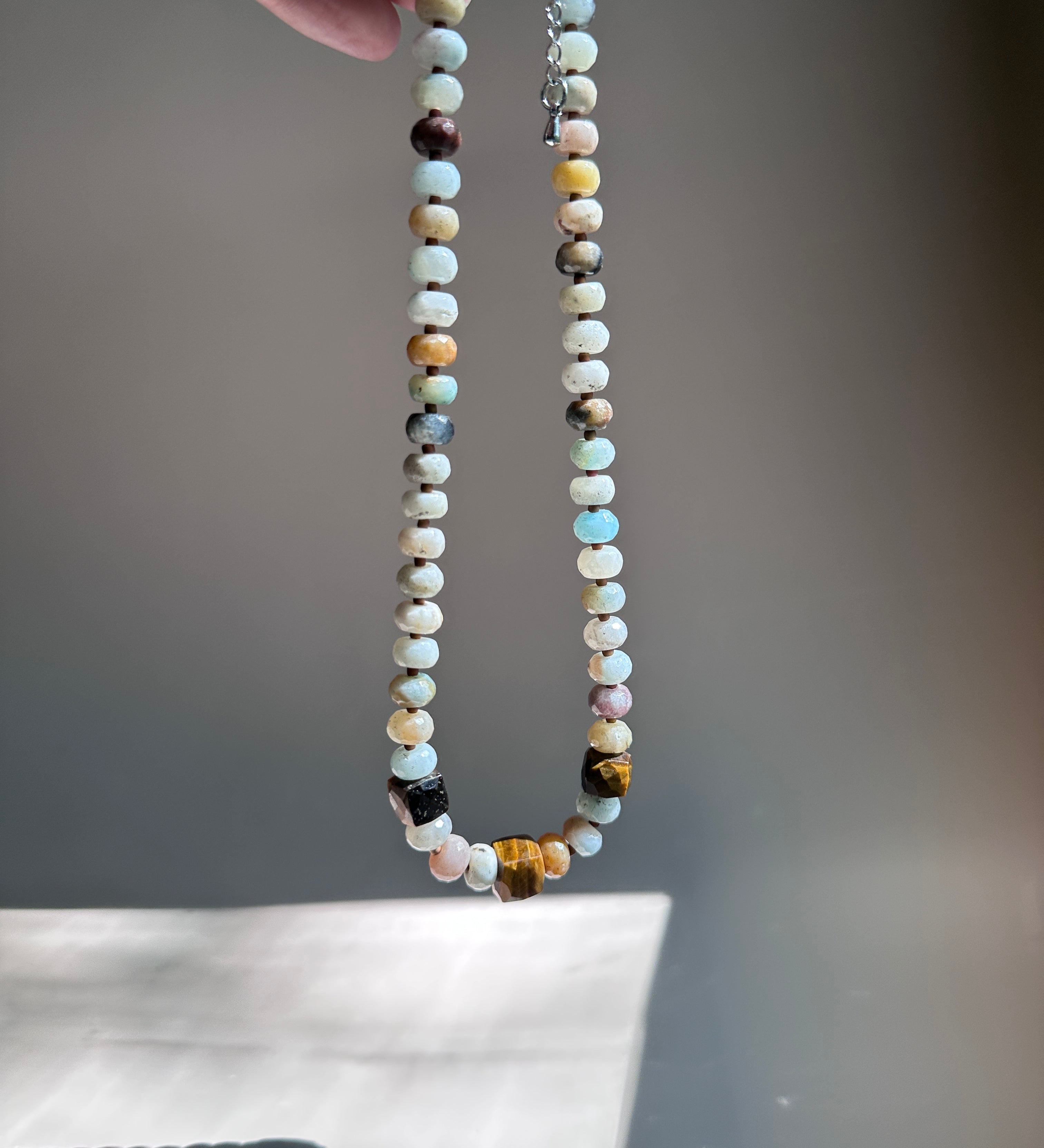A handmade beaded necklace made of multi-colored natural stones, held up by a hand against a neutral gray background.