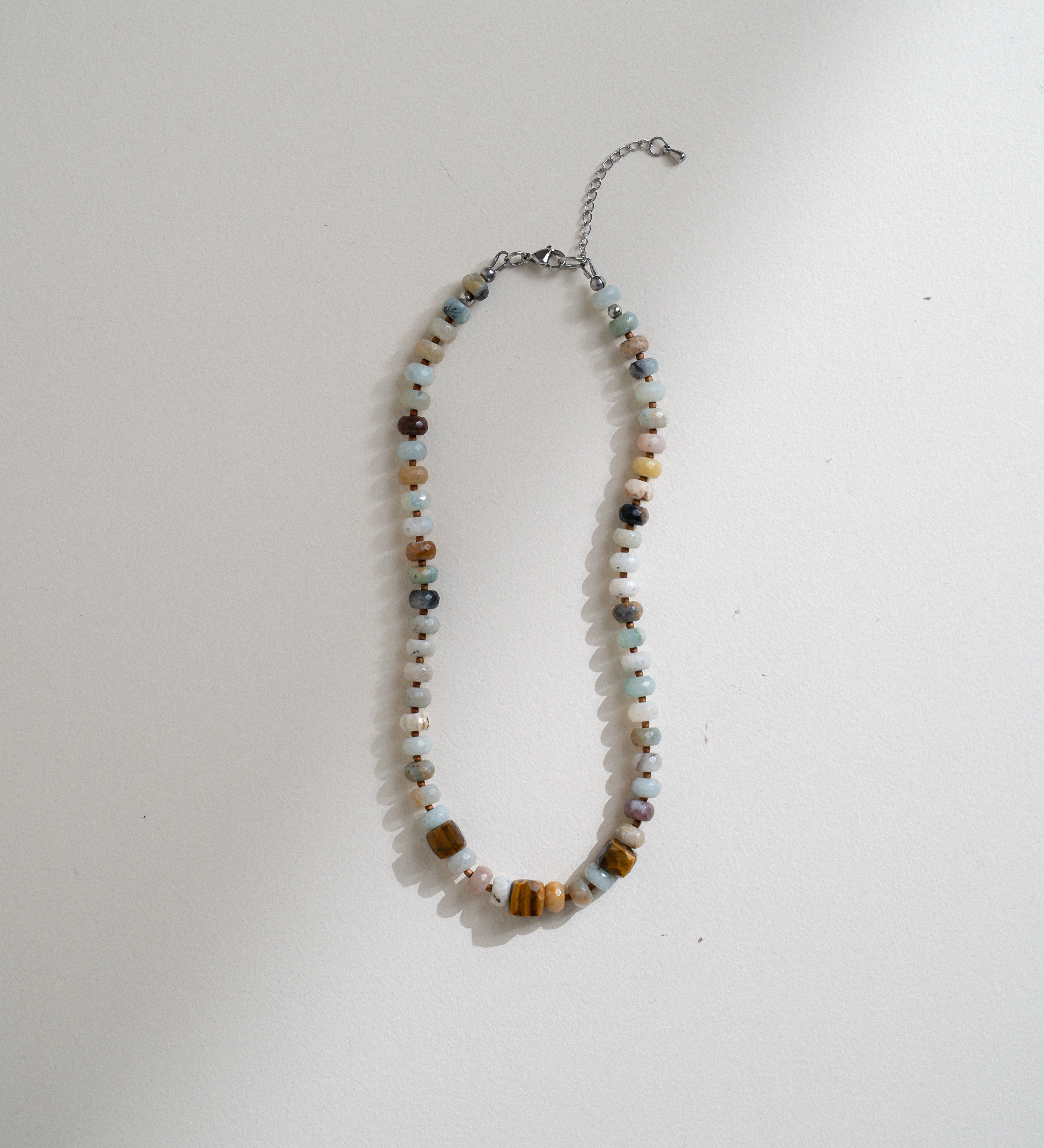 A beaded necklace with a mix of colorful round and square beads in brown, white, light blue, and orange, featuring a silver chain extender, laid flat on a white surface.