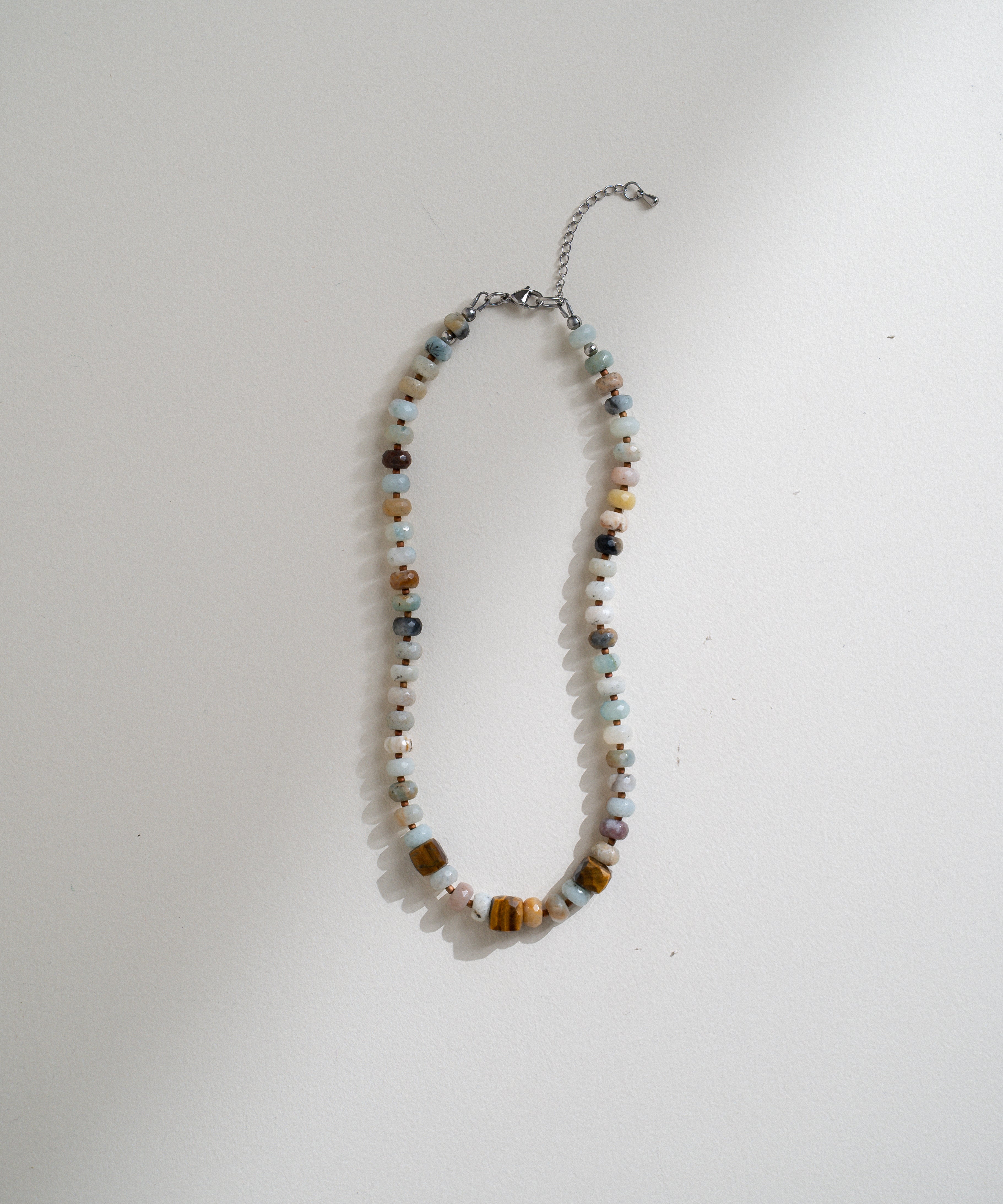 A beaded necklace with a mix of colorful round and square beads in brown, white, light blue, and orange, featuring a silver chain extender, laid flat on a white surface.