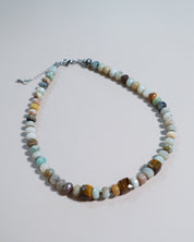 A beaded necklace made of polished multi-colored gemstone beads in shades of brown, green, blue, and white, with a silver-tone clasp and extender chain, displayed on a light gray background.
