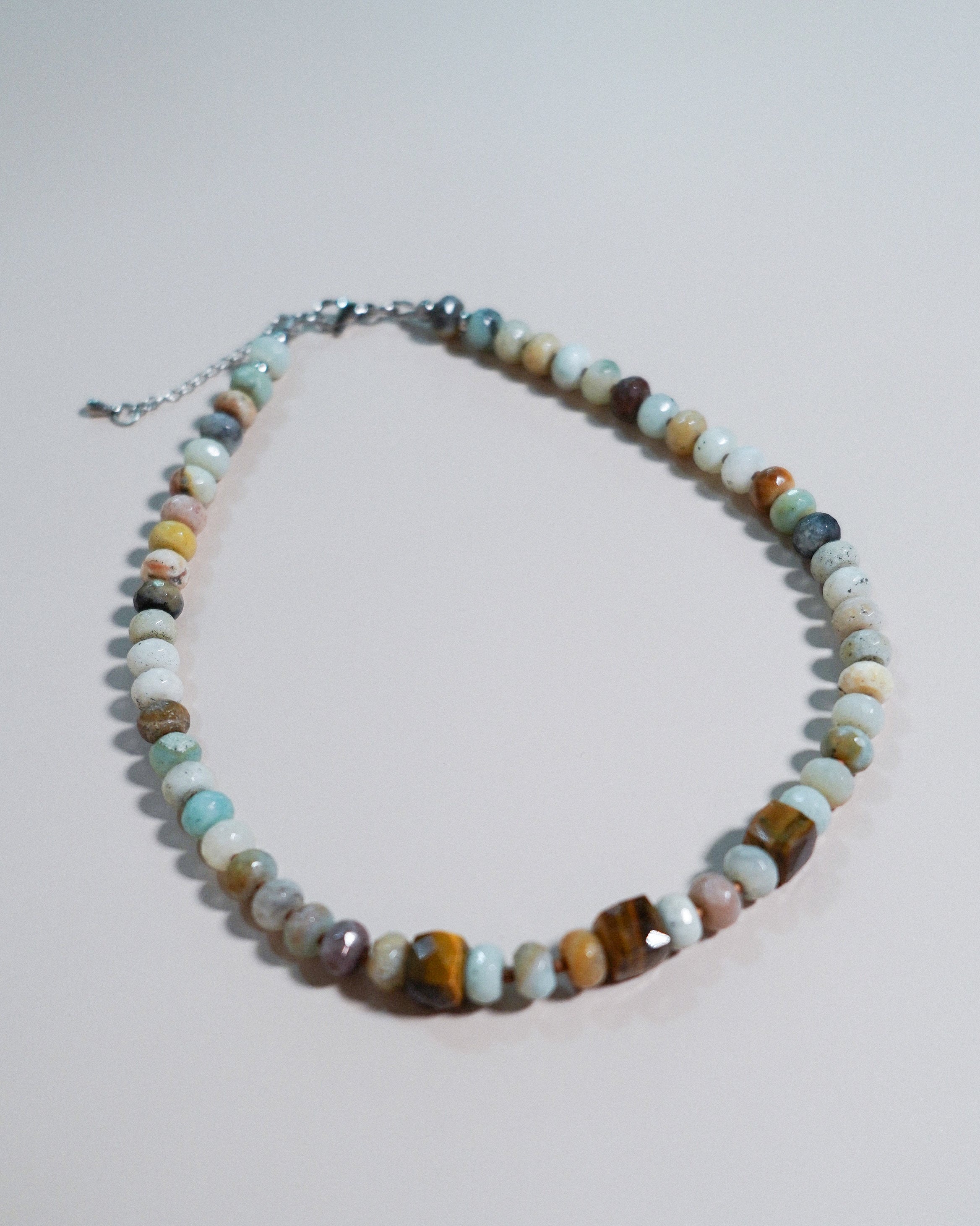 A beaded necklace made of polished multi-colored gemstone beads in shades of brown, green, blue, and white, with a silver-tone clasp and extender chain, displayed on a light gray background.