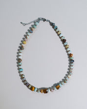A beaded necklace featuring a mix of polished turquoise, brown, yellow, and white stones, accented with small silver beads and a silver chain extender, displayed on a light gray background.