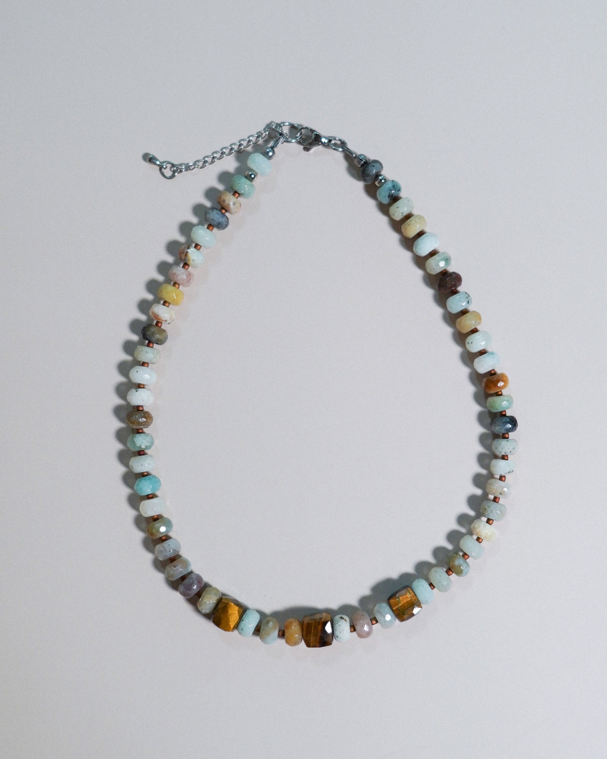 A beaded necklace featuring a mix of polished turquoise, brown, yellow, and white stones, accented with small silver beads and a silver chain extender, displayed on a light gray background.