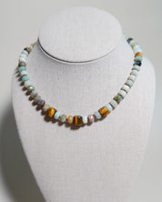 A beaded necklace displayed on a light gray fabric-covered jewelry bust. The necklace features a mix of polished and matte beads in turquoise, brown, orange, and cream colors, with a central cluster of tiger's eye beads.
