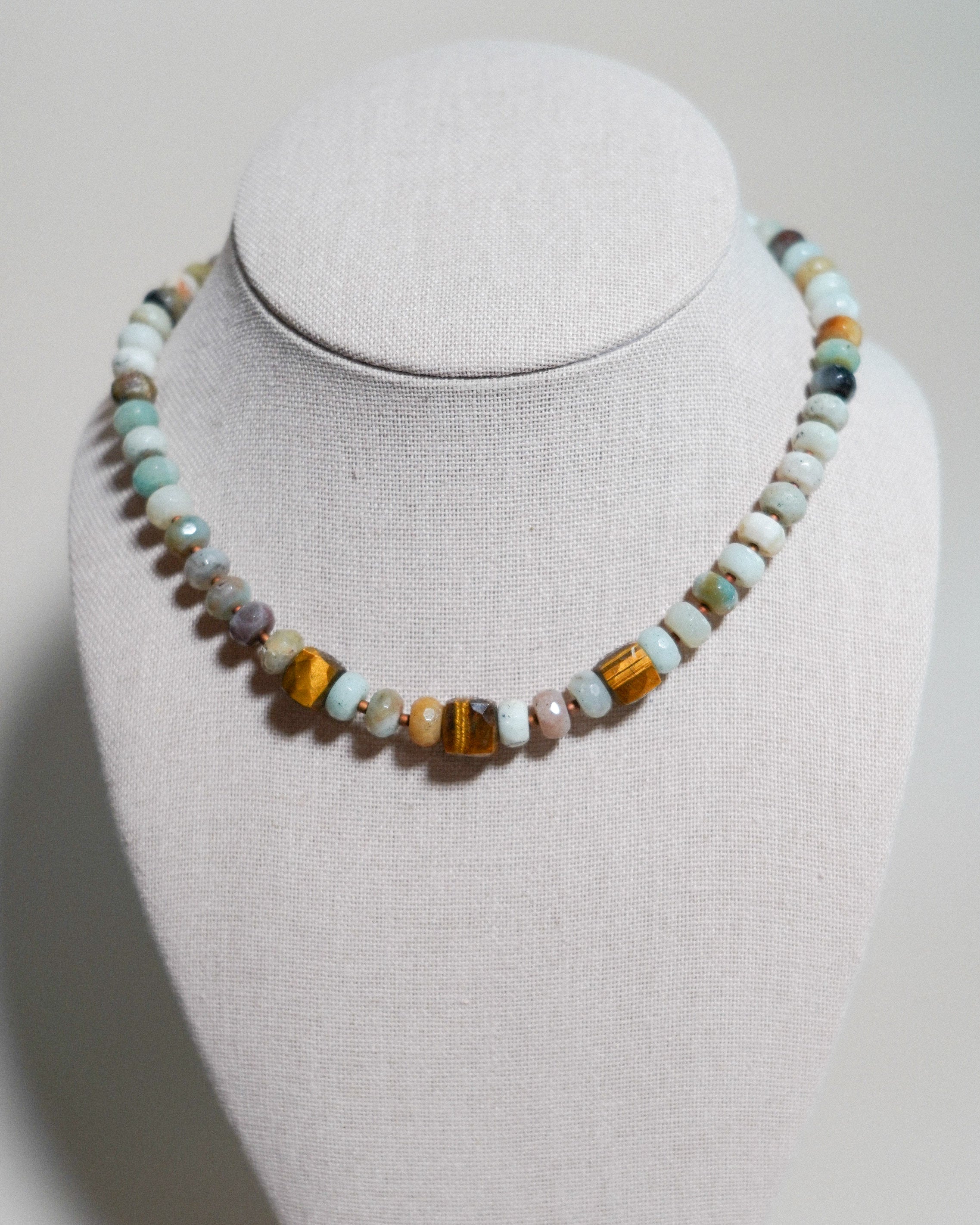 A beaded necklace displayed on a light gray fabric-covered jewelry bust. The necklace features a mix of polished and matte beads in turquoise, brown, orange, and cream colors, with a central cluster of tiger's eye beads.