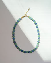 A beaded necklace made of small round beads in shades of blue, green, and teal, with a gold clasp, displayed on a white surface with diagonal shadows.