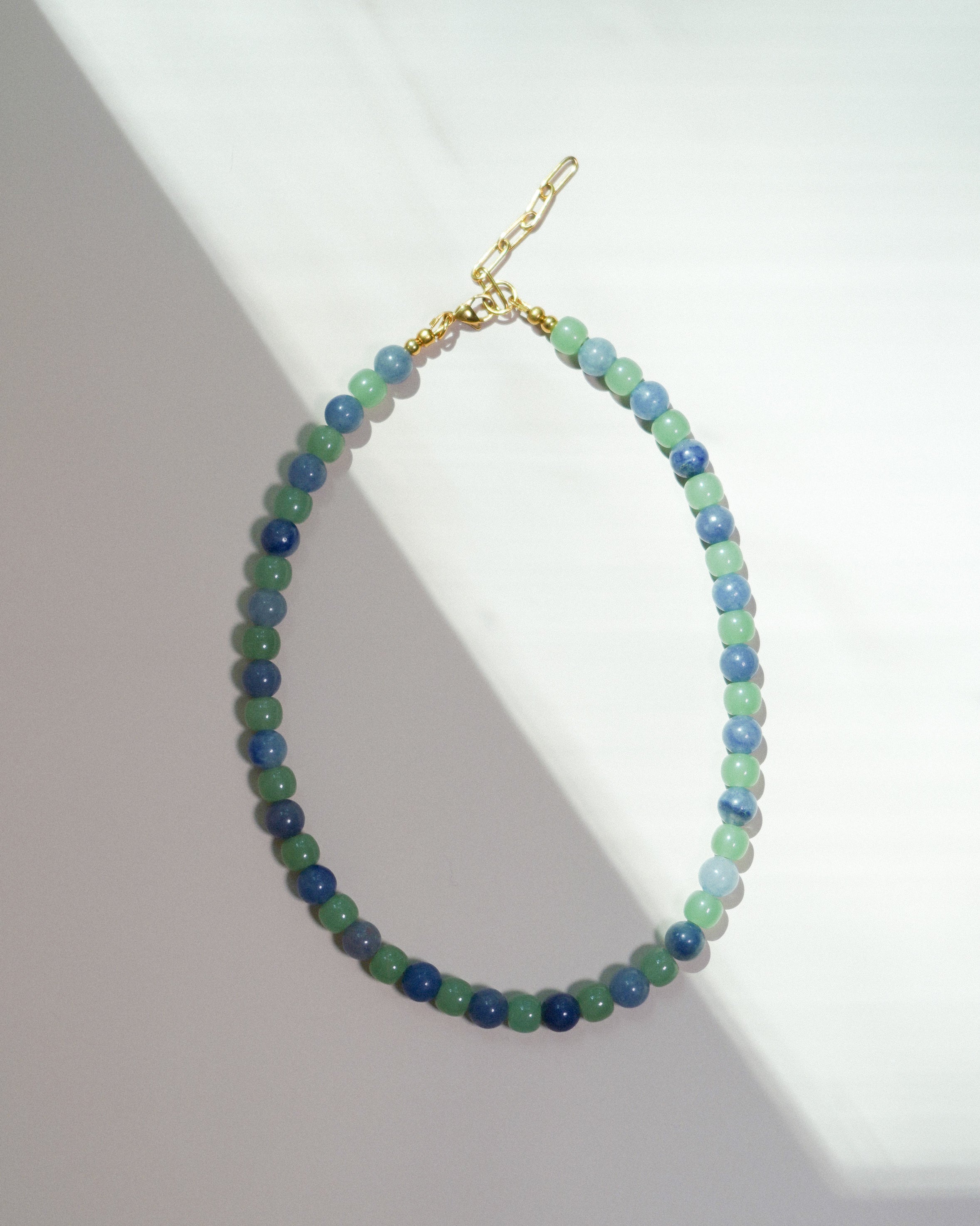 A beaded necklace made of small round beads in shades of blue, green, and teal, with a gold clasp, displayed on a white surface with diagonal shadows.