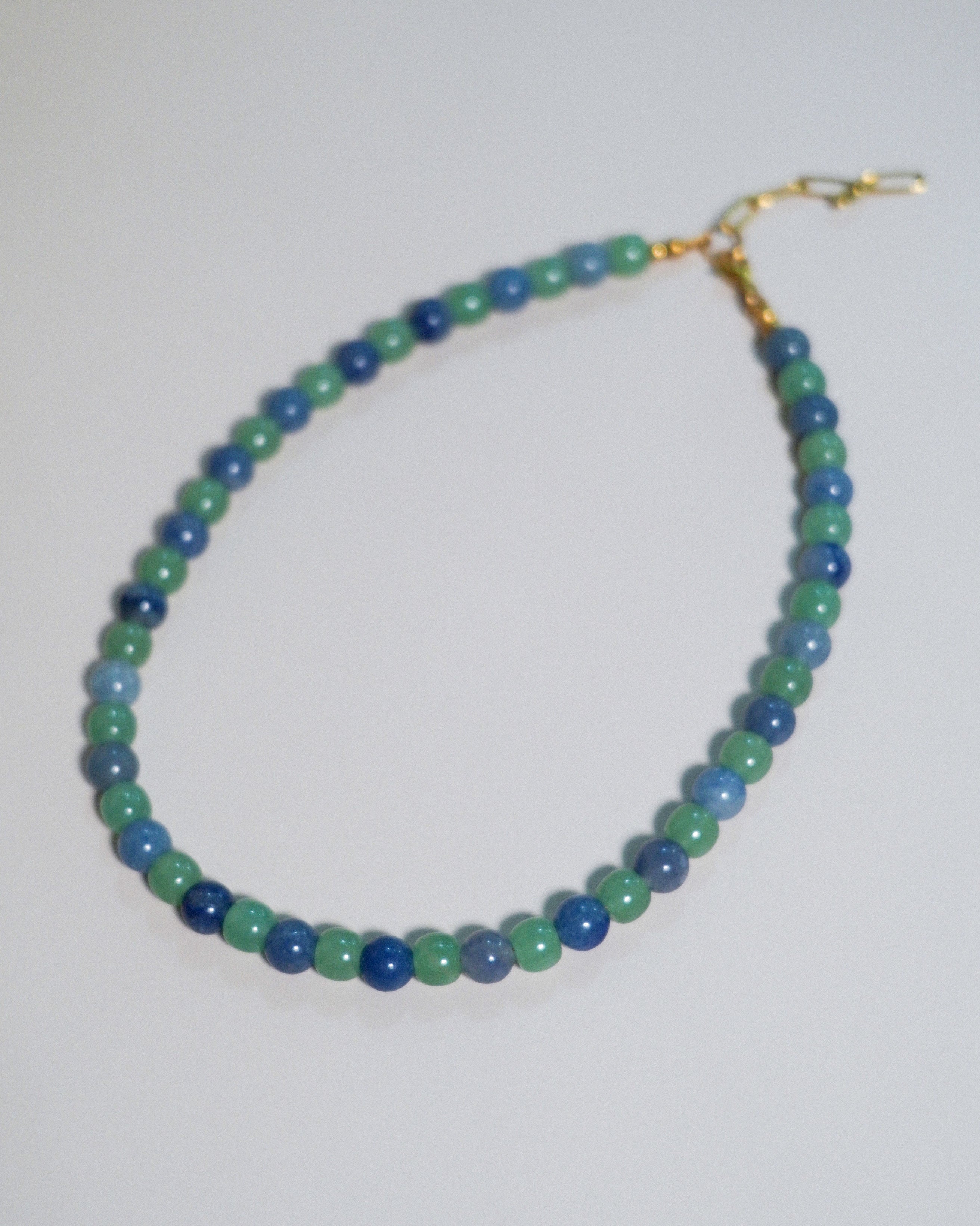 A beaded necklace made of alternating green and blue beads, finished with a gold clasp, shown on a plain light background.