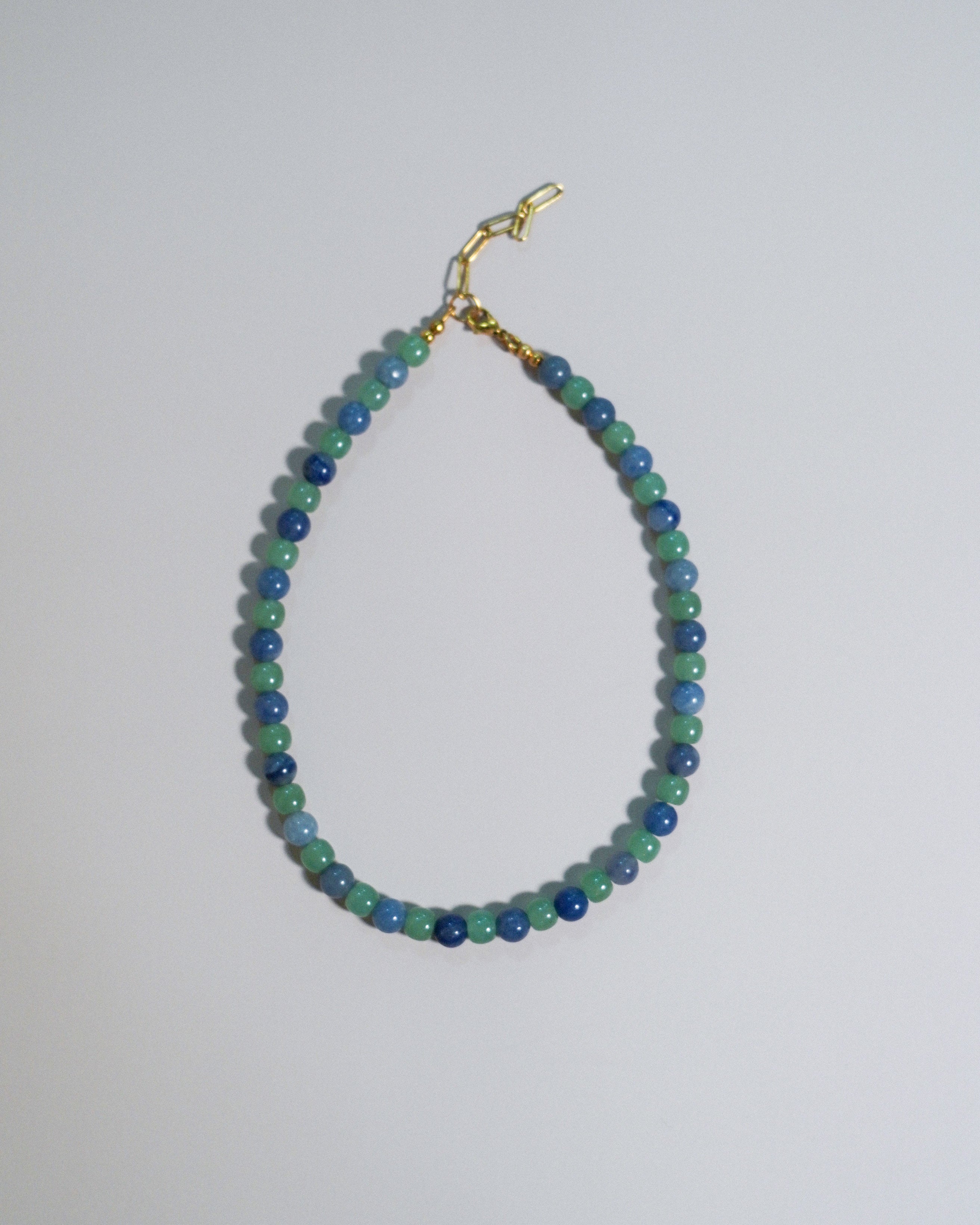 A beaded necklace made of alternating blue and green beads, finished with a gold clasp, displayed on a plain light gray background.