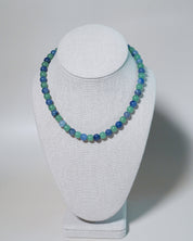A beaded necklace with alternating light green and blue beads, displayed on a white fabric jewelry bust against a light gray background.