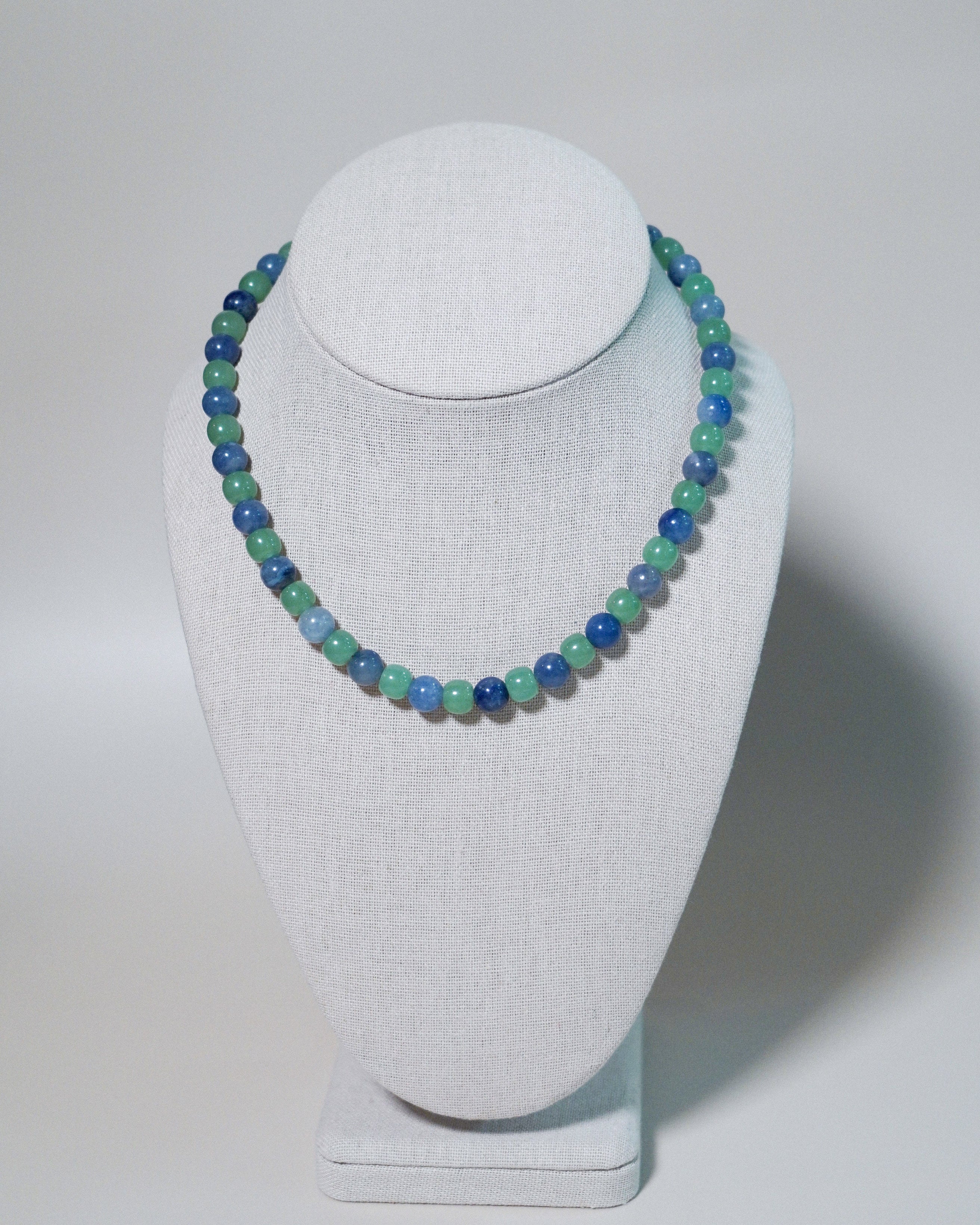 A beaded necklace with alternating light green and blue beads, displayed on a white fabric jewelry bust against a light gray background.
