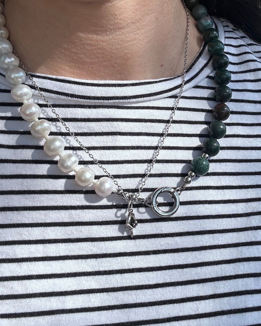 A close-up of a person wearing layered necklaces over a black and white striped shirt. The necklaces include a strand of white pearls with a silver charm, a dark green beaded necklace, and a silver chain with a large open ring and small pendant.