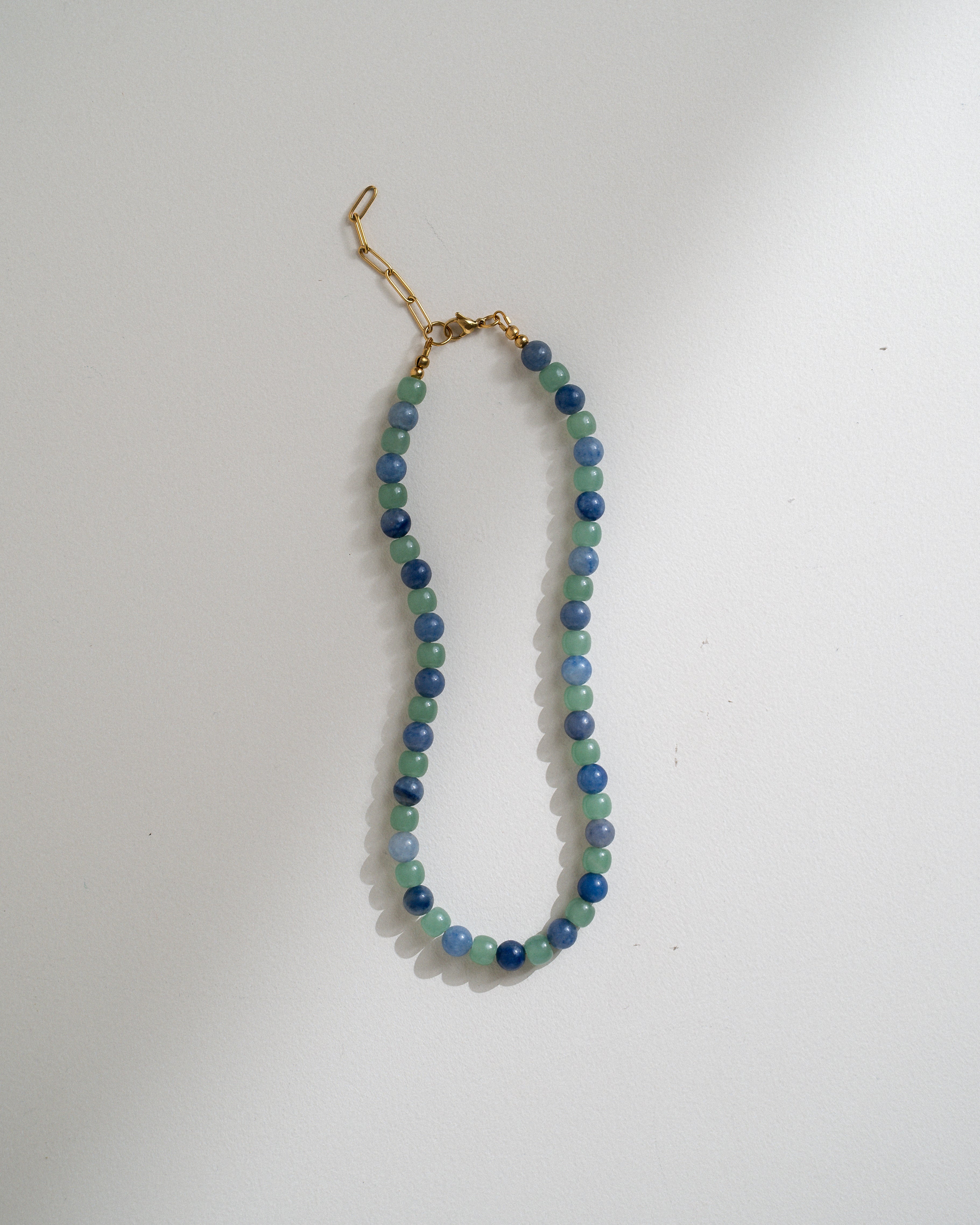 A beaded necklace made of alternating blue and green beads, finished with a gold clasp, displayed on a plain white surface.