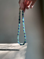 A beaded necklace with alternating light green and dark blue beads, held by a hand against a warm, sunlit gray background.