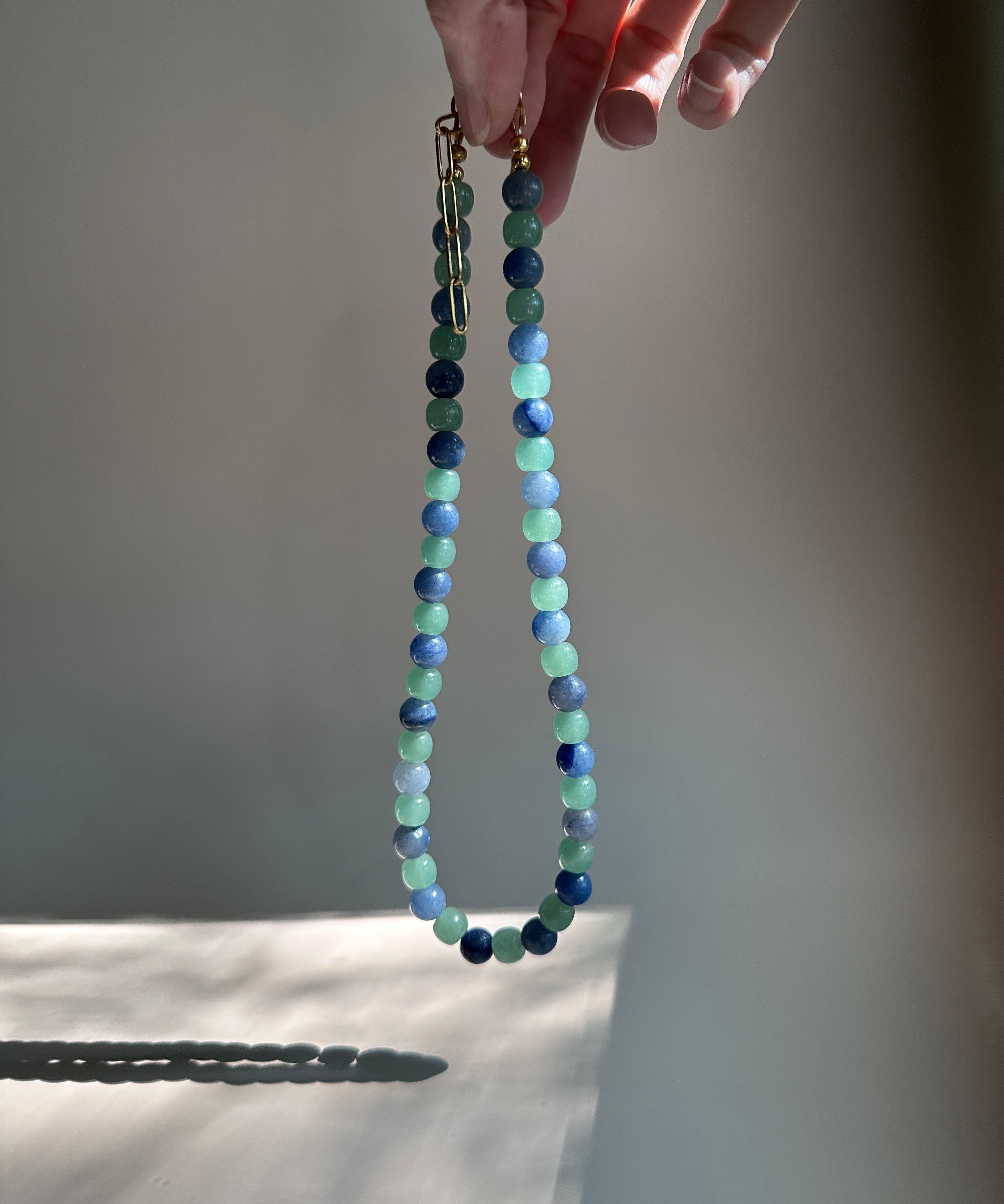 A beaded necklace with alternating light green and dark blue beads, held by a hand against a warm, sunlit gray background.