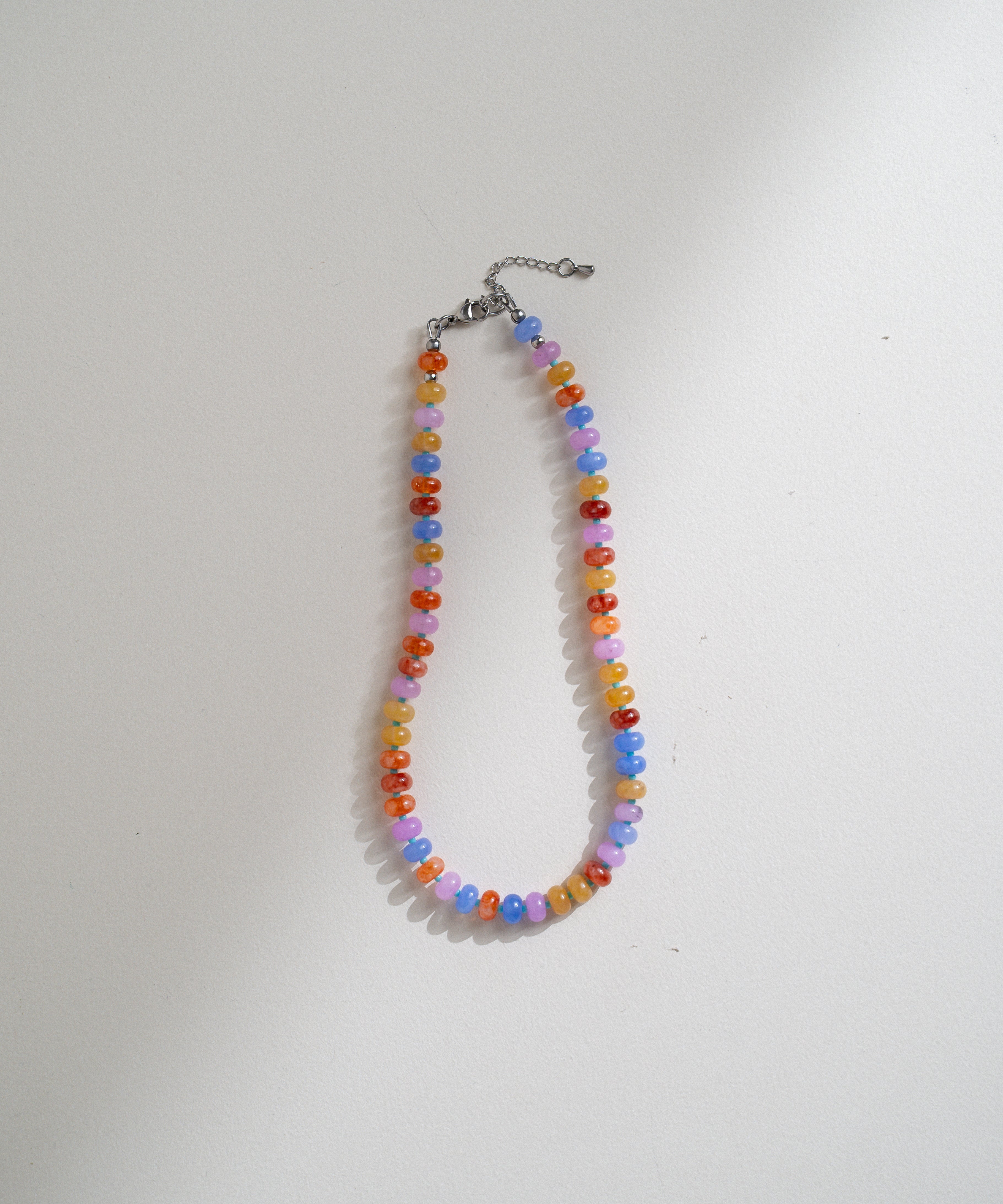 A beaded necklace made of small round beads in pastel rainbow colors, hanging from a silver-toned chain clasp on a plain white background.