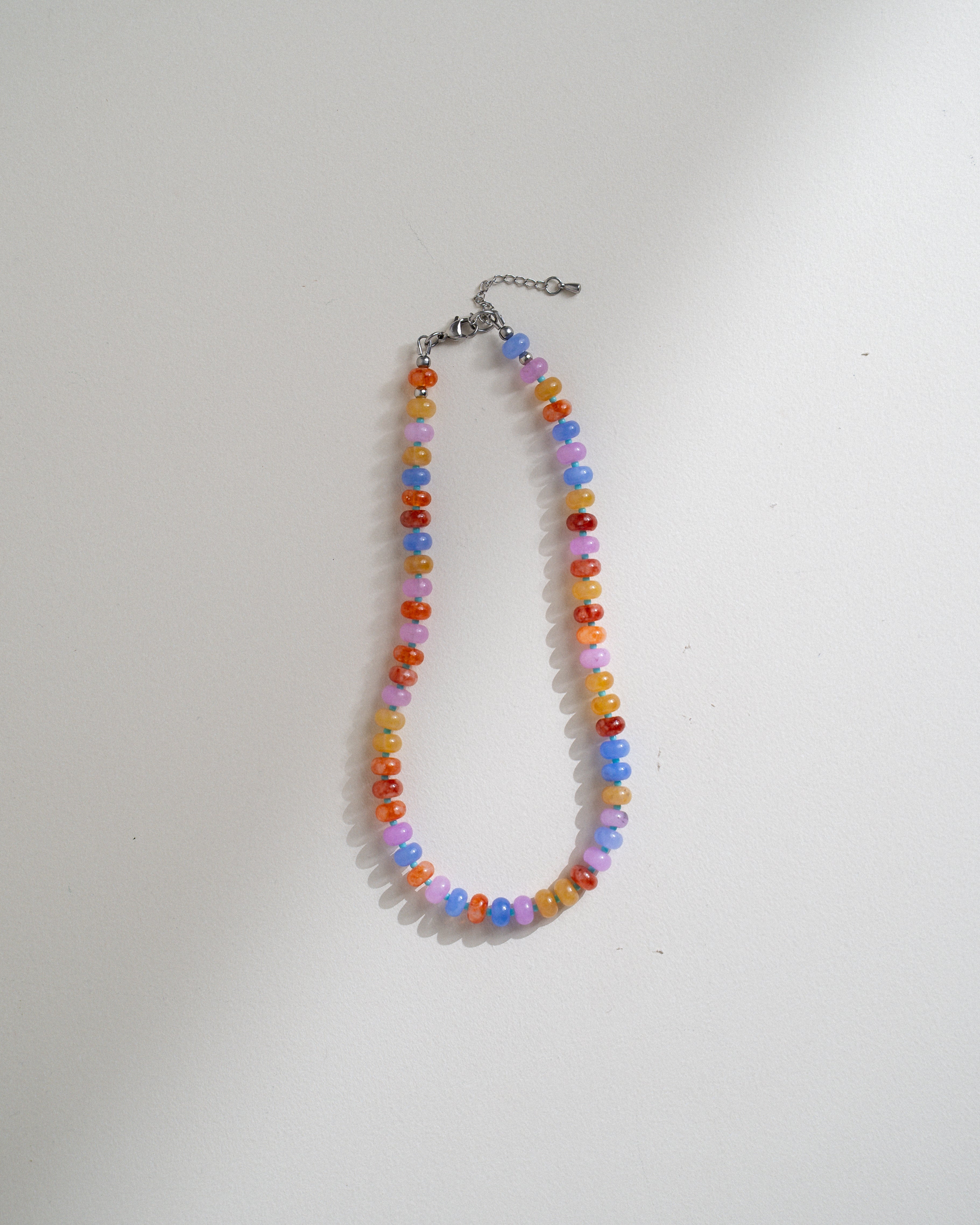 A beaded necklace made of small round beads in pastel rainbow colors, hanging from a silver-toned chain clasp on a plain white background.