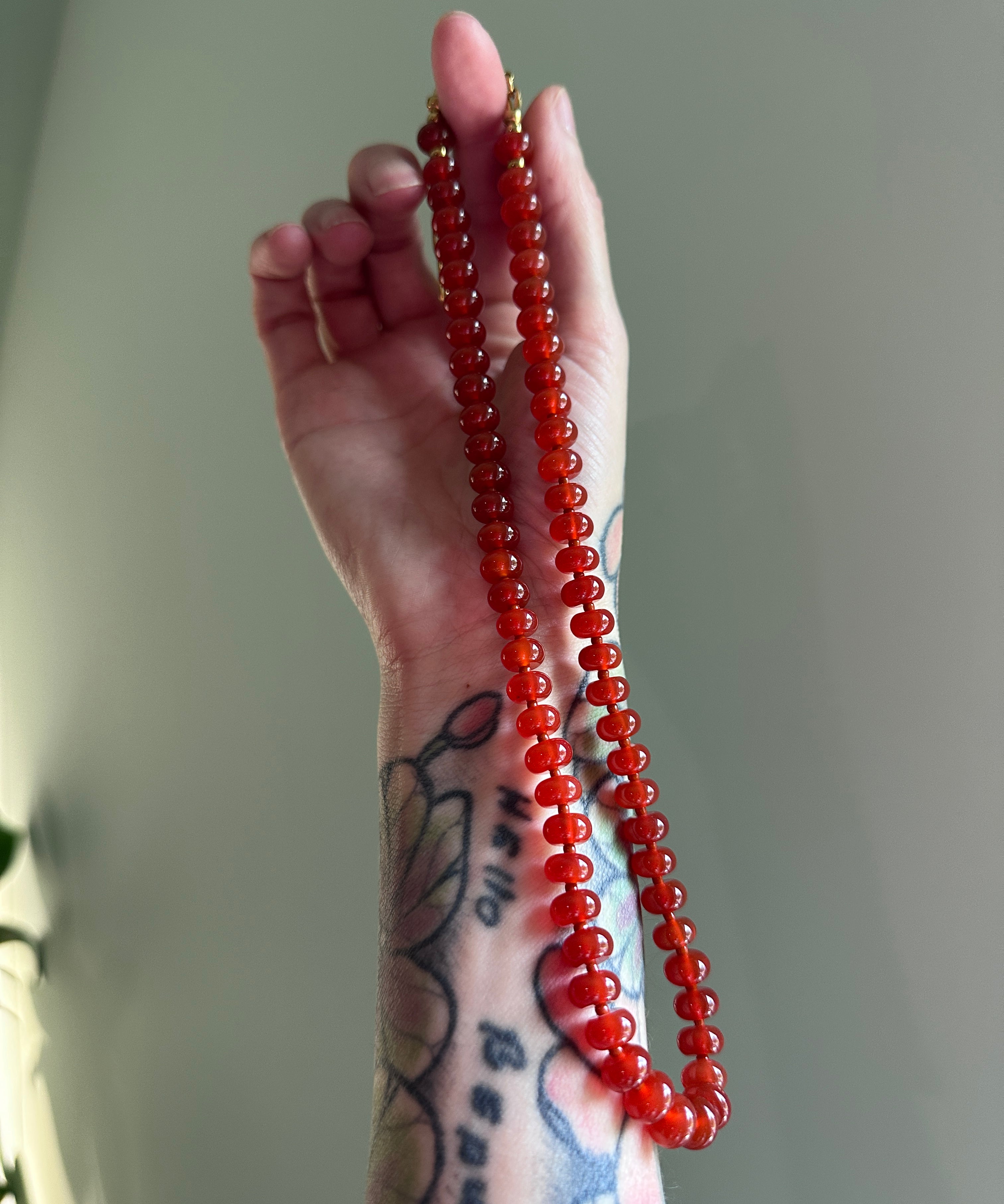 A hand with visible tattoos holds up a long strand of glossy red carnelian beads, showing their translucent orange-red color and smooth rounded texture against a soft gray background.