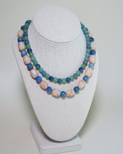 A double-strand beaded necklace displayed on a white fabric jewelry bust. The necklace features alternating strands of polished light pink stone beads and medium blue stone beads, with smaller green beads woven throughout.