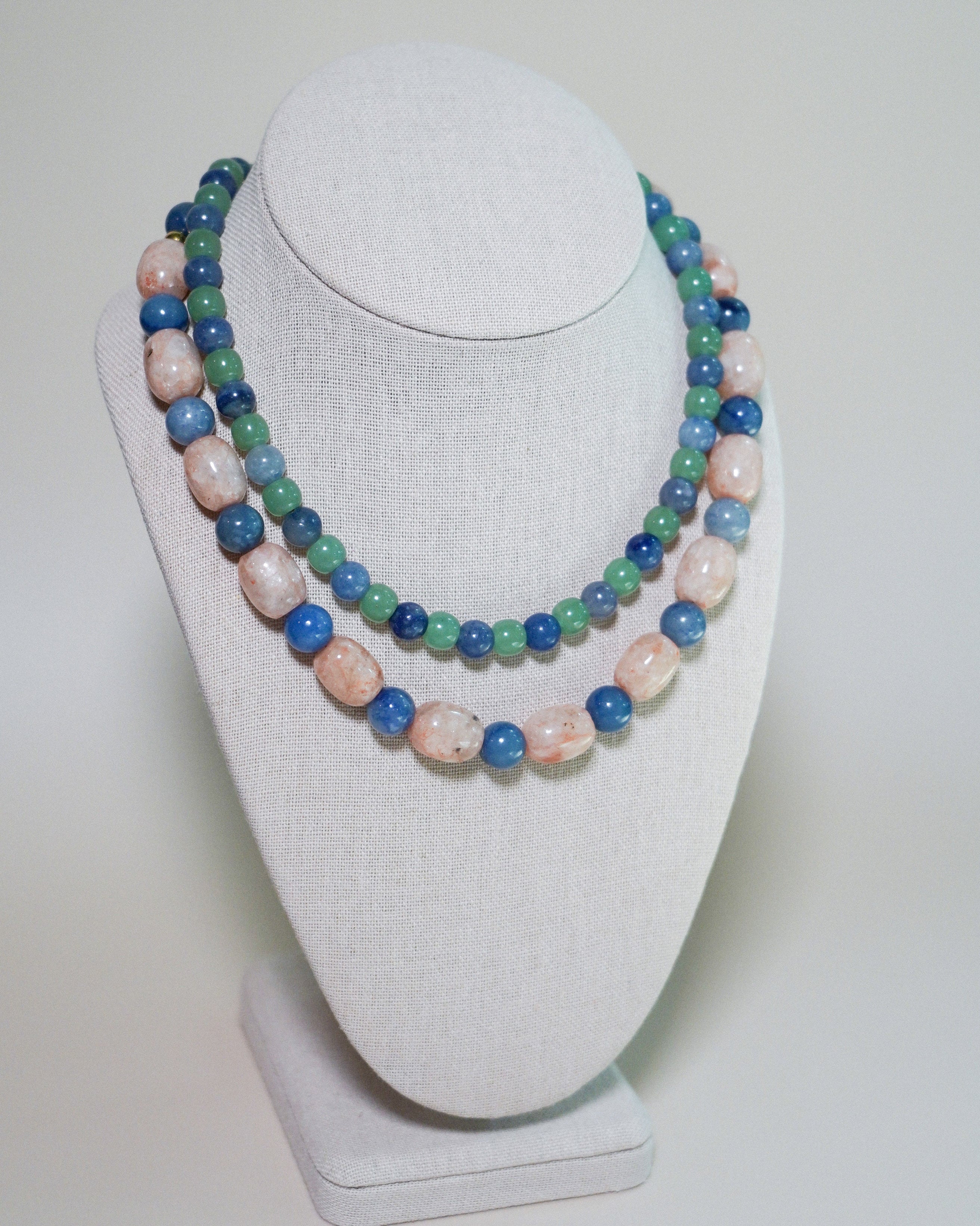A double-strand beaded necklace displayed on a white fabric jewelry bust. The necklace features alternating strands of polished light pink stone beads and medium blue stone beads, with smaller green beads woven throughout.