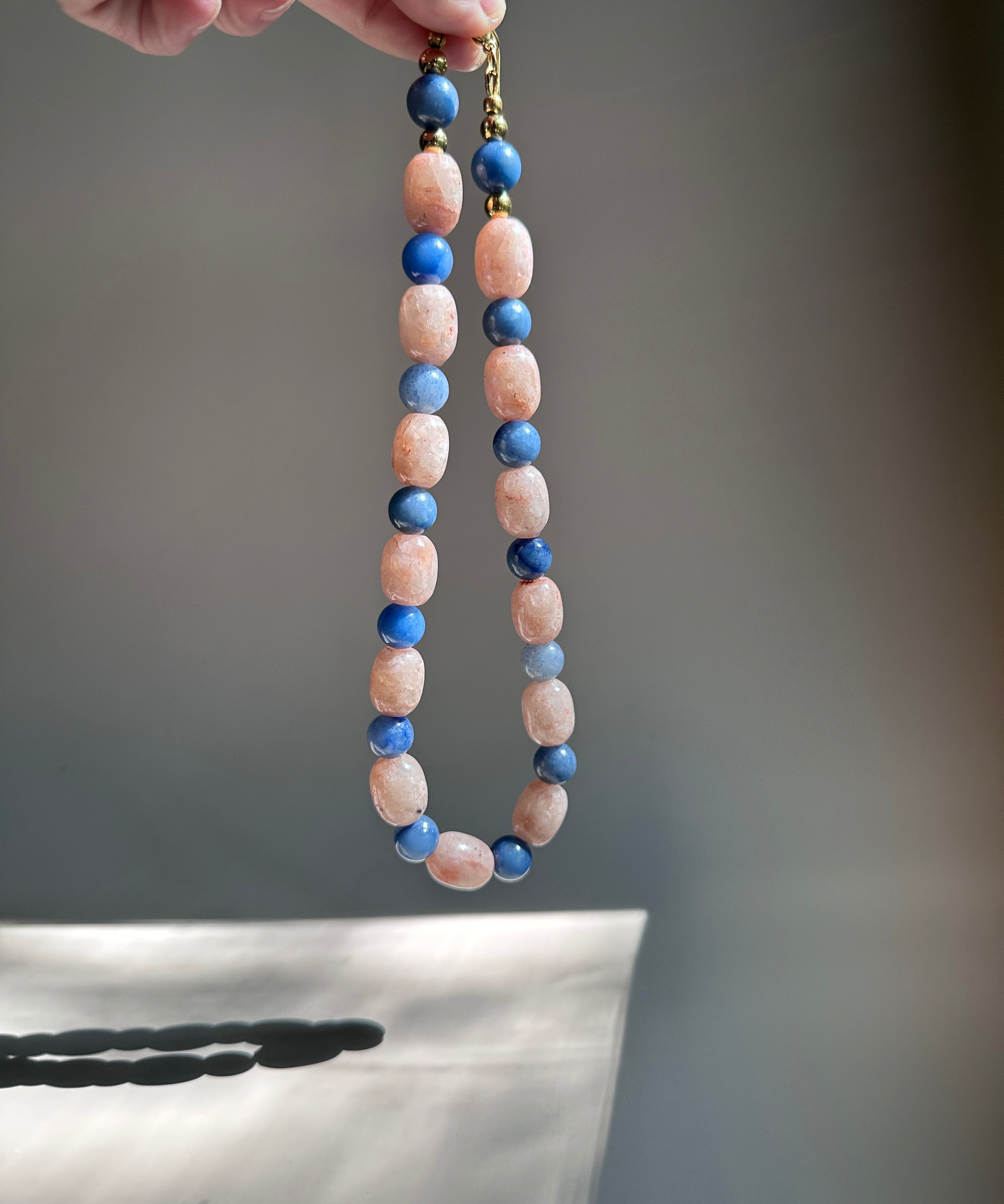 A beaded necklace with alternating light pink and dark blue beads, held by a hand against a soft gray background.