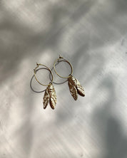 A pair of gold-toned hoop earrings with dangling textured leaf charms, displayed on a light gray surface with soft leaf shadows.