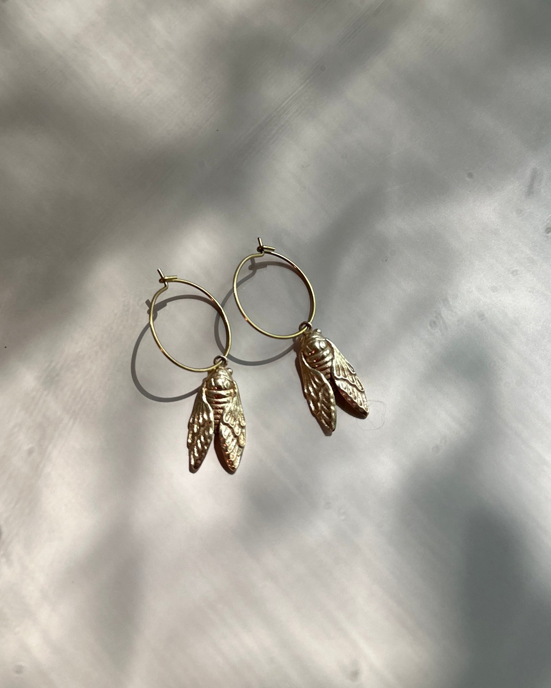 A pair of gold-toned hoop earrings with dangling textured leaf charms, displayed on a light gray surface with soft leaf shadows.