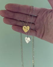 Hand holding two heart-shaped necklaces with gold and silver chains against a light green background