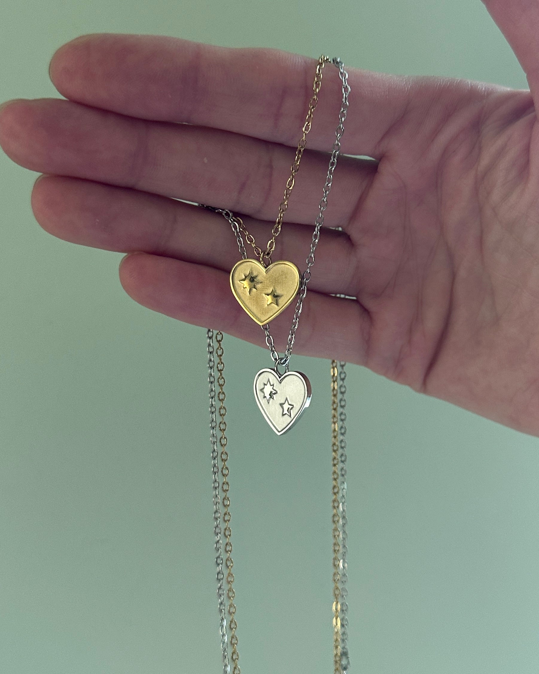 Hand holding two heart-shaped necklaces with gold and silver chains against a light green background