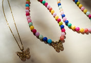 Three beaded necklaces with butterfly pendants displayed on a light beige surface. One necklace has a gold chain and gold butterfly pendant.