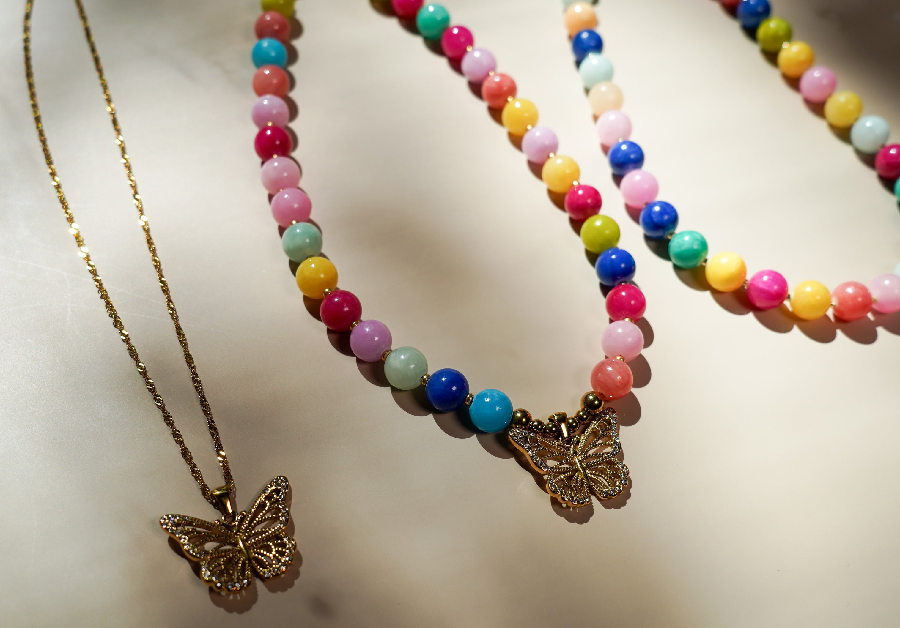 Three beaded necklaces with butterfly pendants displayed on a light beige surface. One necklace has a gold chain and gold butterfly pendant.